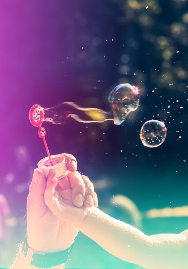 A man holding a child's hand whilst they blow bubbles with a bubble blower in the sun.