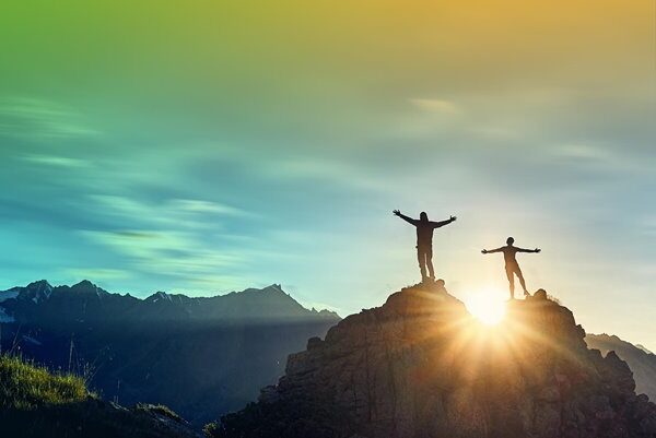 Two climbers with their arms spread wide on the top of a mountain with a sunrise.