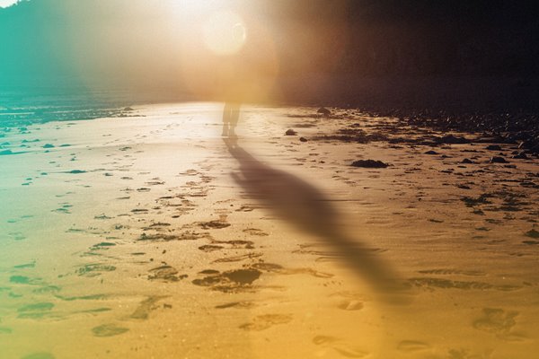 Person with a long shadow walking on a sandy beach towards the sun leaving footprints
